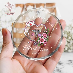 May include: A circular faceted glass ornament featuring the text "OFFICIALLY DISCHARGED" and "Retired Nurse -2026-". The design includes a heart, a nurse cap, a stethoscope, a caduceus symbol, the name "Amanda", and pink floral accents.