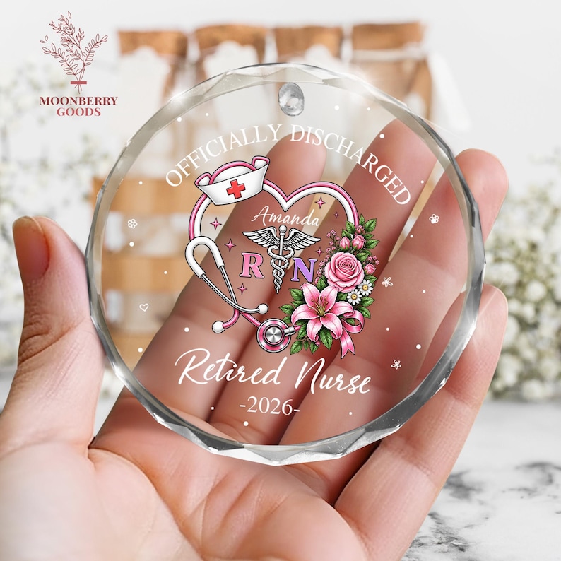 May include: A circular faceted glass ornament featuring the text "OFFICIALLY DISCHARGED" and "Retired Nurse -2026-". The design includes a heart, a nurse cap, a stethoscope, a caduceus symbol, the name "Amanda", and pink floral accents.