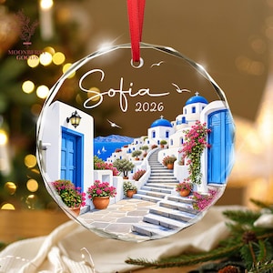 May include: A circular crystal ornament featuring a printed scene of a Greek island with white buildings, blue domes, and vibrant pink flowers. The text Sofia 2026 is printed in white script. A red ribbon is attached to the top for hanging.