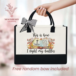 May include: A canvas tote bag with black handles and a black bottom. The bag features the text "This is how I fight my battles" with a floral design and a cross. A black and white striped bow is attached to the handle.