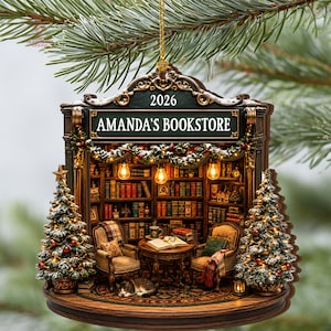 May include: A decorative hanging ornament shaped like a bookstore storefront with the text 2026 and AMANDA'S BOOKSTORE. The scene features two armchairs, a table with an open book, a sleeping cat, bookshelves, and two Christmas trees with lights.