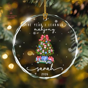 May include: A round, faceted clear glass ornament featuring the text "THE YEAR I LEARNED mahjong." above a Christmas tree decorated with mahjong tiles, pink flowers, and blue ornaments. The name "sarah" and "2026" are printed below.