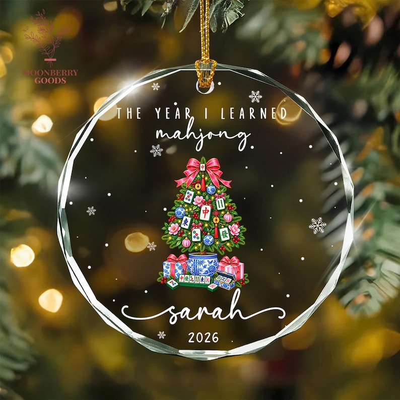 May include: A round, faceted clear glass ornament featuring the text "THE YEAR I LEARNED mahjong." above a Christmas tree decorated with mahjong tiles, pink flowers, and blue ornaments. The name "sarah" and "2026" are printed below.