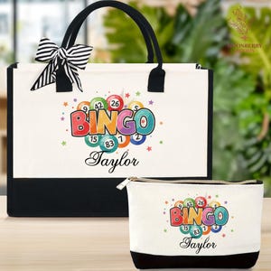 May include: A white tote bag and matching cosmetic bag with a black base and handles. Both bags feature a colorful "BINGO" graphic with bingo balls and the name "Taylor". A black and white striped bow adorns the tote bag.