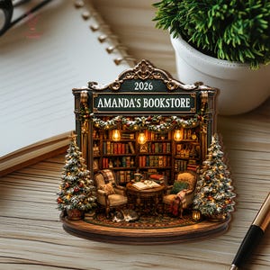 May include: A decorative ornament shaped like a bookstore interior with the text 2026 and AMANDA'S BOOKSTORE. It features two armchairs, a small table with an open book, a sleeping cat, two Christmas trees, and shelves filled with books.