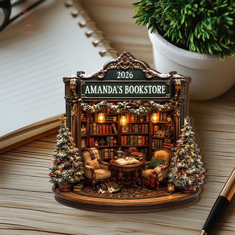 May include: A decorative ornament shaped like a bookstore interior with the text 2026 and AMANDA'S BOOKSTORE. It features two armchairs, a small table with an open book, a sleeping cat, two Christmas trees, and shelves filled with books.