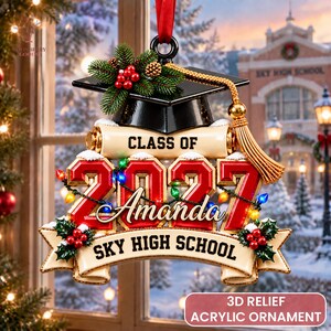 May include: A 3D relief acrylic ornament featuring a black graduation cap topped with pine branches and red berries. Below, a scroll reads "CLASS OF 2027" with the name "Amanda" and "SKY HIGH SCHOOL" on a banner, accented by colourful string lights.