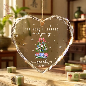 May include: A heart-shaped clear crystal ornament featuring the text THE YEAR I LEARNED mahjong, a decorated Christmas tree with mahjong tiles, and the name Sarah 2026. The ornament is suspended by a gold cord and decorated with white snowflakes.