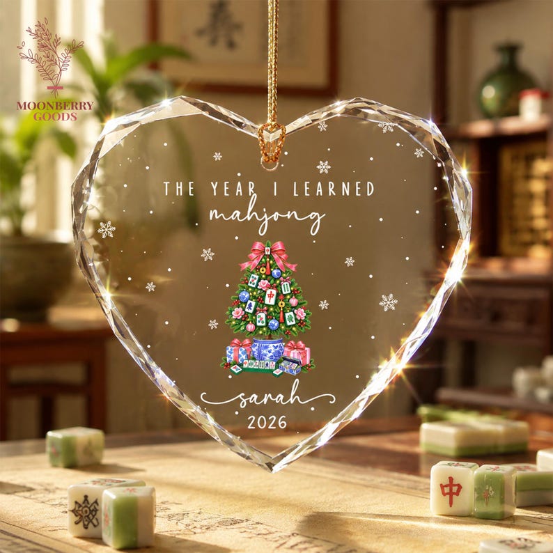 May include: A heart-shaped clear crystal ornament featuring the text THE YEAR I LEARNED mahjong, a decorated Christmas tree with mahjong tiles, and the name Sarah 2026. The ornament is suspended by a gold cord and decorated with white snowflakes.