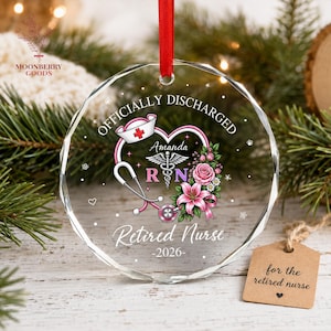 May include: A clear, faceted, circular glass ornament hanging from a red ribbon. It features a printed design with a nurse cap, a stethoscope, a caduceus, pink flowers, and the text "OFFICIALLY DISCHARGED", "Amanda", "RN", "Retired Nurse", and "2026".