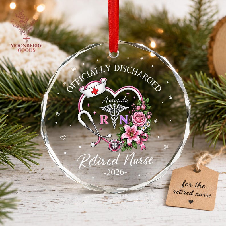 May include: A clear, faceted, circular glass ornament hanging from a red ribbon. It features a printed design with a nurse cap, a stethoscope, a caduceus, pink flowers, and the text "OFFICIALLY DISCHARGED", "Amanda", "RN", "Retired Nurse", and "2026".