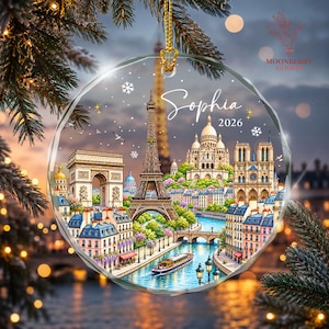 May include: A round, clear glass ornament featuring a detailed illustration of the Paris skyline, including the Eiffel Tower, Arc de Triomphe, and Sacré-Cœur. The text Sophia and 2026 are printed in white script and sans-serif font on the ornament.