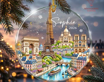May include: A round, clear glass ornament featuring a detailed illustration of the Paris skyline, including the Eiffel Tower, Arc de Triomphe, and Sacré-Cœur. The text Sophia and 2026 are printed in white script and sans-serif font on the ornament.