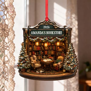 May include: A decorative hanging ornament shaped like a bookstore interior with a red ribbon. The sign reads 2026 AMANDA'S BOOKSTORE. It features bookshelves, two armchairs, a small table, a sleeping cat, and two Christmas trees with lights.