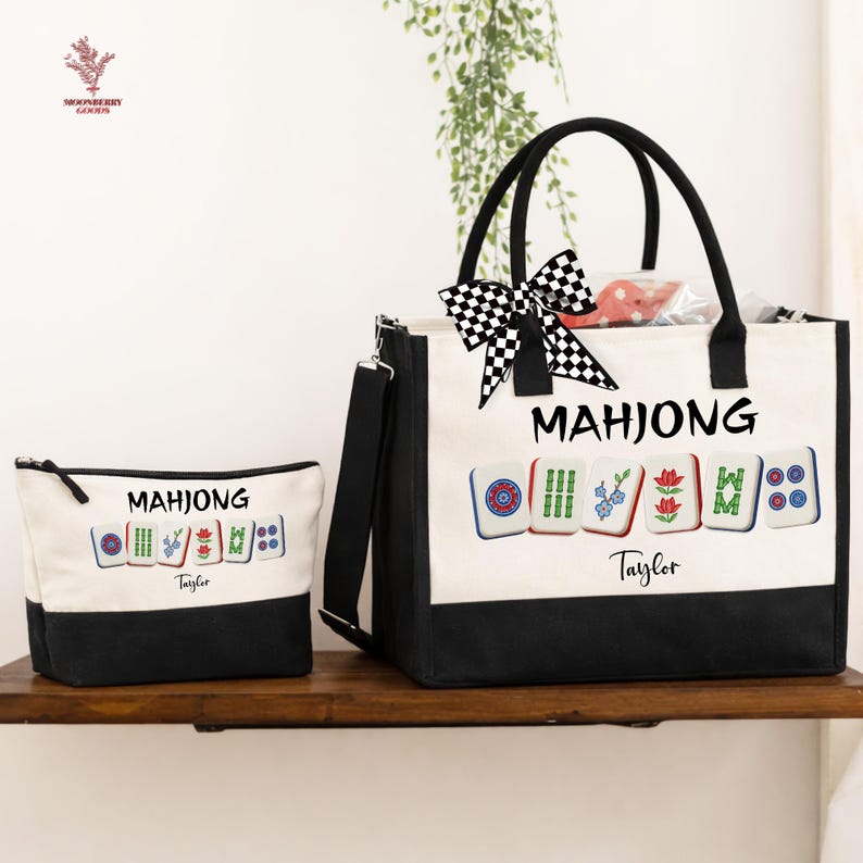May include: A canvas tote bag and matching pouch set. The bags are primarily off-white with black accents and feature the word "MAHJONG" and colorful tile illustrations. A black and white checkered bow adorns the tote bag.