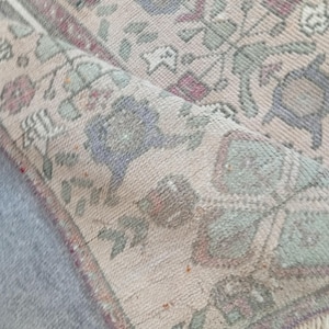 May include: A rolled-up, vintage-style rug with a beige background and floral patterns in shades of blue, green, and red. The rug has a fringed edge and a distressed appearance, suggesting age and use. The design features stylized flowers and leaves.