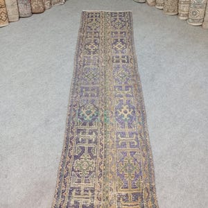 Puede incluir: Una alfombra de pasillo larga y estrecha con un patrón geométrico repetitivo en tonos morados, verdes apagados y beige. El diseño consta de motivos rectangulares interconectados y bordes, rematado con flecos cortos en ambos extremos de la pieza.