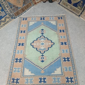 May include: A rectangular, hand-woven rug with a central geometric design in light blue, peach, and navy blue. The rug has a light green center and a peach border with navy blue geometric shapes. The rug is placed on a gray surface.