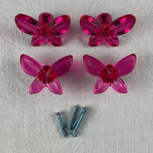 May include: Four translucent pink butterfly-shaped cabinet knobs. Each knob has a central body and four wings. Three silver screws are positioned below the knobs. The knobs are likely made of acrylic or glass.