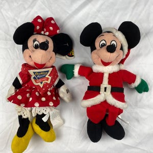 K&ouml;nnte beinhalten: Zwei Pl&uuml;schpuppen von Disney-Charakteren. Eine ist Minnie Mouse in einem roten gepunkteten Kleid und einer Schleife. Die andere ist Mickey Mouse in einem Weihnachtsmann-Outfit. Beide Puppen haben schwarze Ohren und l&auml;chelnde Gesichter.