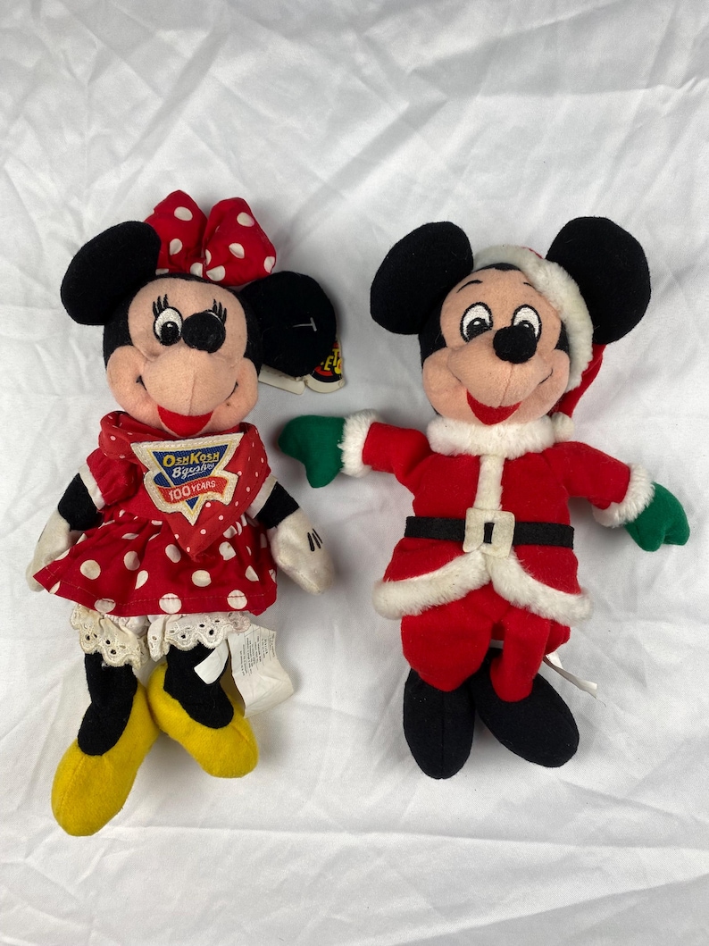 K&ouml;nnte beinhalten: Zwei Pl&uuml;schpuppen von Disney-Charakteren. Eine ist Minnie Mouse in einem roten gepunkteten Kleid und einer Schleife. Die andere ist Mickey Mouse in einem Weihnachtsmann-Outfit. Beide Puppen haben schwarze Ohren und l&auml;chelnde Gesichter.