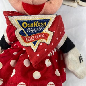 K&ouml;nnte beinhalten: Minnie Mouse Pl&uuml;schpuppe mit rotem Kleid mit wei&szlig;en Punkten. Die Puppe hat schwarze Handschuhe und ein Etikett mit der Aufschrift "OshKosh B'gosh 100 Years". Das Gesicht der Puppe ist rosa mit einem roten L&auml;cheln.