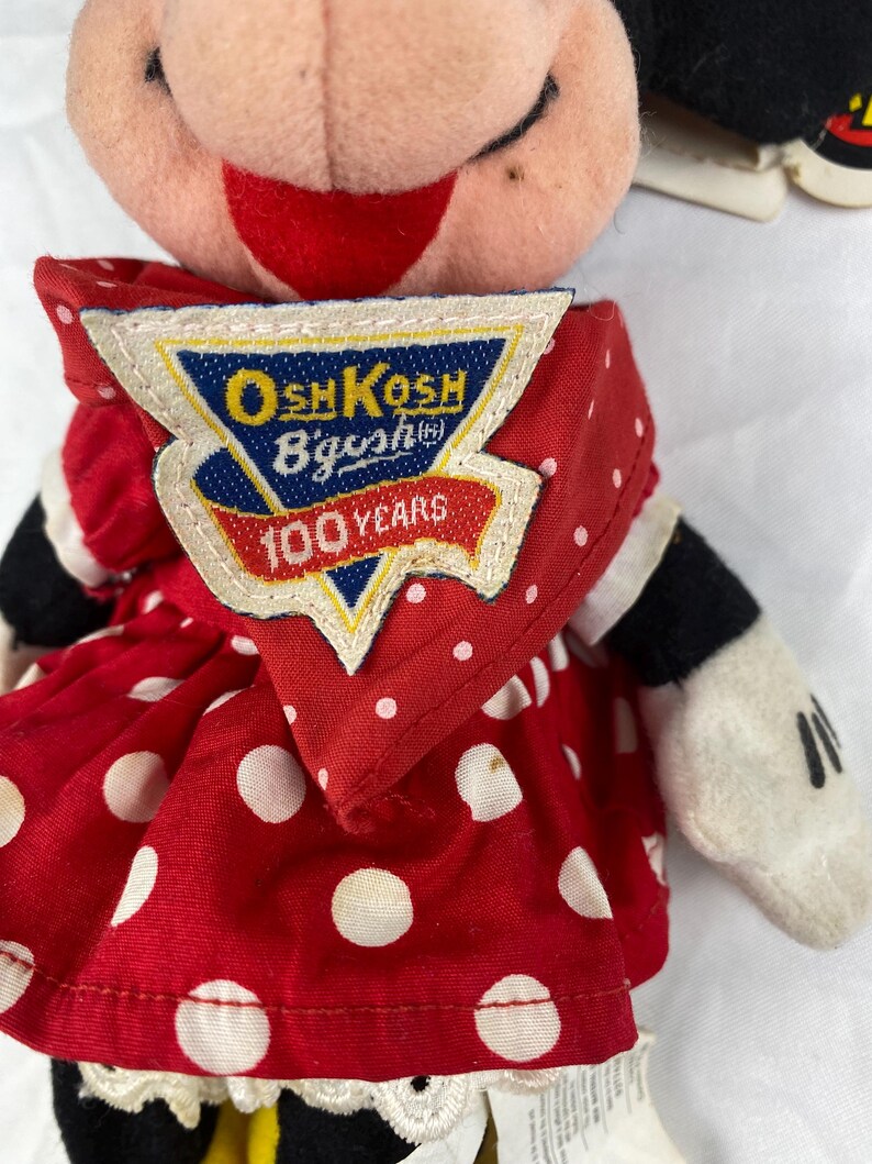 K&ouml;nnte beinhalten: Minnie Mouse Pl&uuml;schpuppe mit rotem Kleid mit wei&szlig;en Punkten. Die Puppe hat schwarze Handschuhe und ein Etikett mit der Aufschrift "OshKosh B'gosh 100 Years". Das Gesicht der Puppe ist rosa mit einem roten L&auml;cheln.