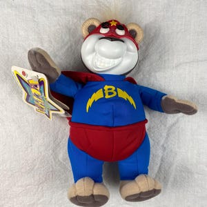 May include: A plush toy bear dressed as a superhero. The bear is wearing a blue and yellow suit with a red cape and a red mask with a yellow star. The bear has a white face with a big smile and a lightning bolt on its chest. The bear is holding its right arm up in the air. The toy is labeled "The Powerpuff Girls".