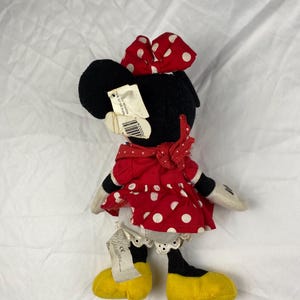 K&ouml;nnte beinhalten: Ein Minnie Mouse Pl&uuml;schtier, mit einem schwarzen Kopf und einer rot-wei&szlig; gepunkteten Schleife. Sie tr&auml;gt ein rotes Kleid mit wei&szlig;en Punkten, wei&szlig;e Handschuhe und gelbe Schuhe. Das Spielzeug hat ein Etikett.