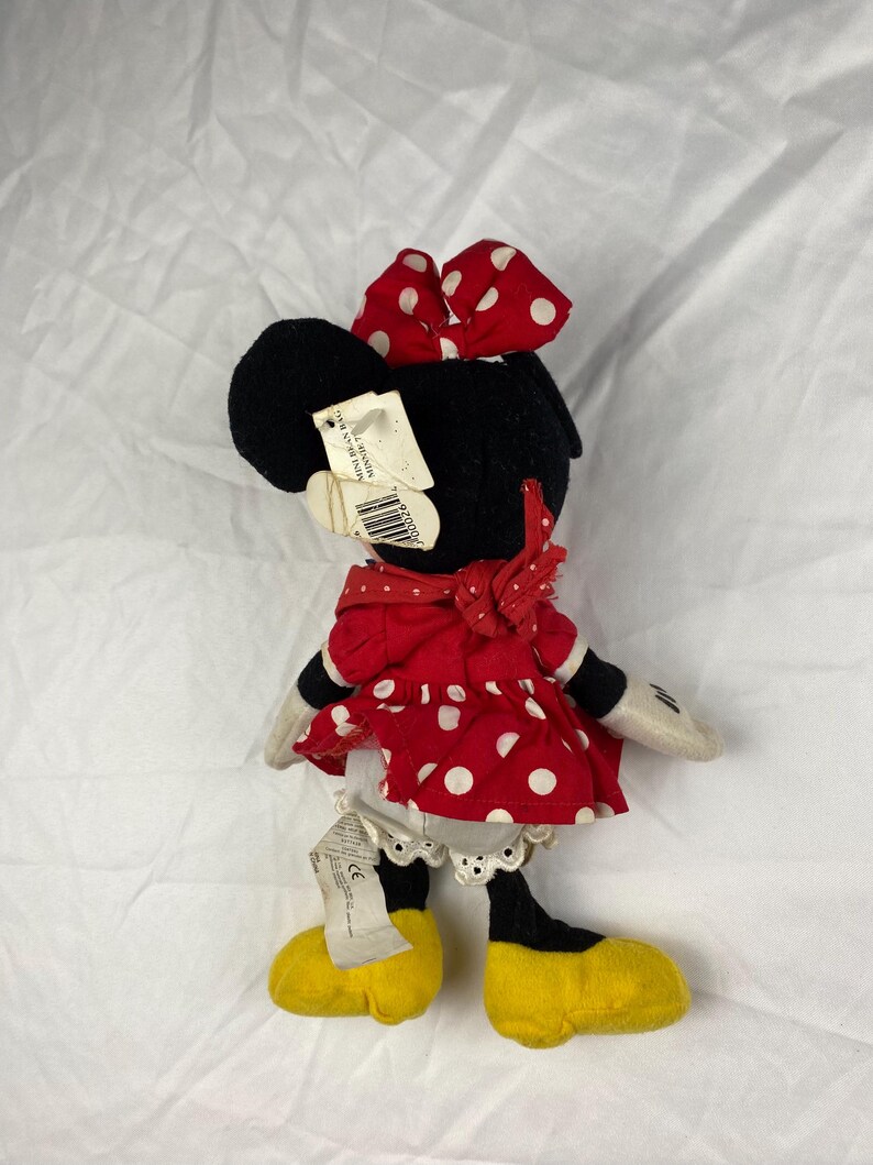 K&ouml;nnte beinhalten: Ein Minnie Mouse Pl&uuml;schtier, mit einem schwarzen Kopf und einer rot-wei&szlig; gepunkteten Schleife. Sie tr&auml;gt ein rotes Kleid mit wei&szlig;en Punkten, wei&szlig;e Handschuhe und gelbe Schuhe. Das Spielzeug hat ein Etikett.