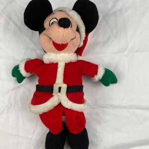 K&ouml;nnte beinhalten: Ein Stofftier von Mickey Mouse als Weihnachtsmann. Die Figur tr&auml;gt einen roten Anzug mit wei&szlig;er Einfassung, einen schwarzen G&uuml;rtel und gr&uuml;ne Handschuhe. Mickey hat eine Weihnachtsm&uuml;tze, schwarze Ohren und Schuhe.