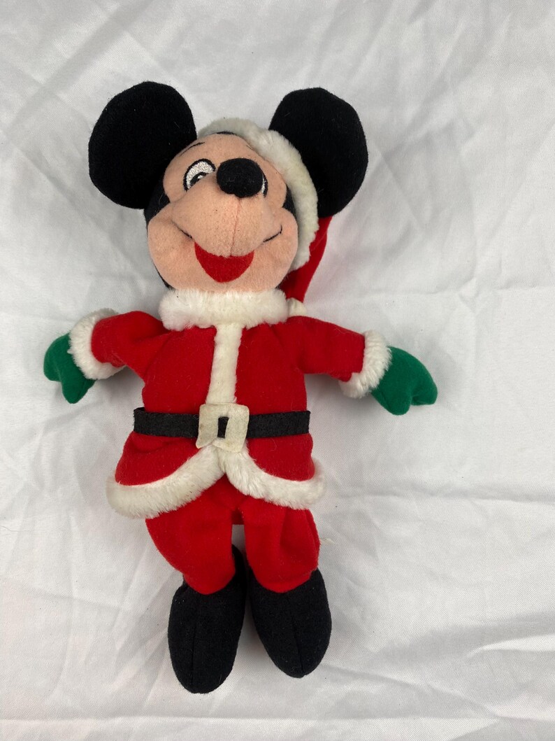 K&ouml;nnte beinhalten: Ein Stofftier von Mickey Mouse als Weihnachtsmann. Die Figur tr&auml;gt einen roten Anzug mit wei&szlig;er Einfassung, einen schwarzen G&uuml;rtel und gr&uuml;ne Handschuhe. Mickey hat eine Weihnachtsm&uuml;tze, schwarze Ohren und Schuhe.