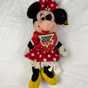 K&ouml;nnte beinhalten: Eine Pl&uuml;schpuppe von Minnie Mouse mit einem roten Kleid mit wei&szlig;en Punkten, einer passenden Schleife und gelben Schuhen. Die Puppe hat schwarze Ohren und einen rot-wei&szlig; gepunkteten Schal. Das Etikett lautet "OshKosh B'gosh 100 Years."