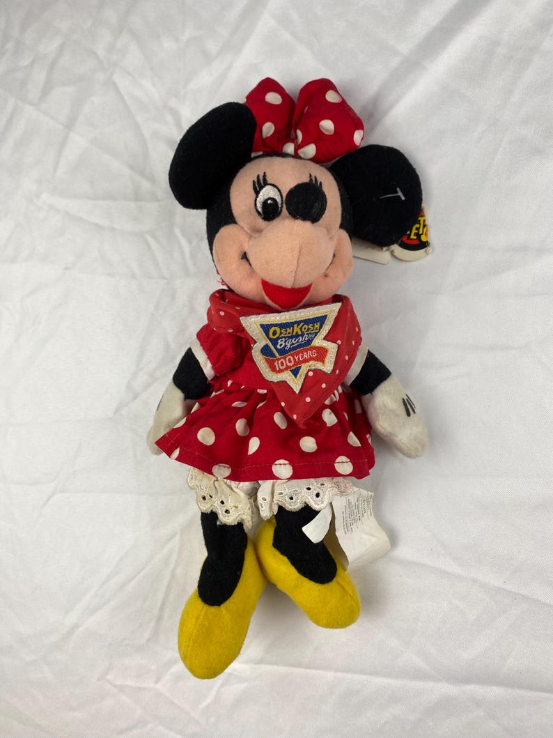 K&ouml;nnte beinhalten: Eine Pl&uuml;schpuppe von Minnie Mouse mit einem roten Kleid mit wei&szlig;en Punkten, einer passenden Schleife und gelben Schuhen. Die Puppe hat schwarze Ohren und einen rot-wei&szlig; gepunkteten Schal. Das Etikett lautet "OshKosh B'gosh 100 Years."