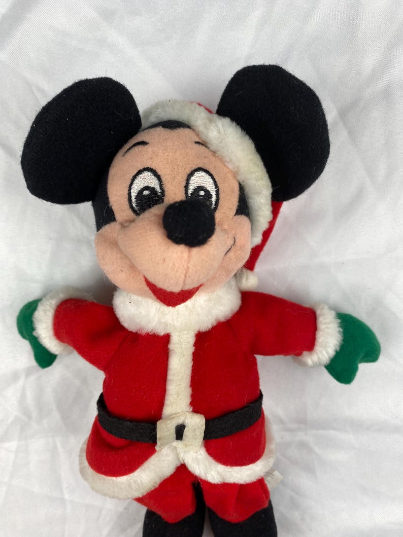 K&ouml;nnte beinhalten: Ein Mickey Mouse Pl&uuml;schtier im Weihnachtsmann-Outfit. Das Spielzeug hat schwarze Ohren, einen roten Weihnachtsmann-Anzug mit wei&szlig;em Besatz, einen schwarzen G&uuml;rtel und eine Weihnachtsm&uuml;tze. Die Handschuhe sind gr&uuml;n.