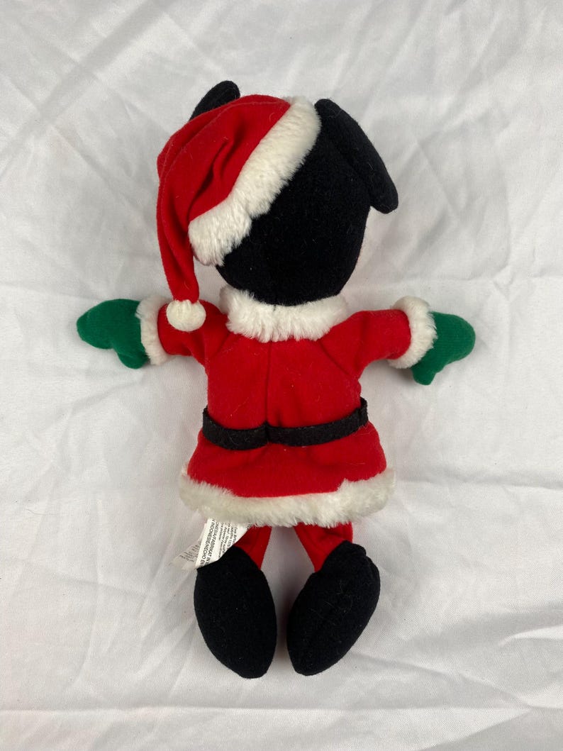 K&ouml;nnte beinhalten: Eine schwarze Pl&uuml;schfigur in einem roten Weihnachtsmann-Anzug mit wei&szlig;er Verzierung, passendem Hut und gr&uuml;nen Handschuhen. Die Figur hat schwarze F&uuml;&szlig;e und ist f&uuml;r die Weihnachtszeit gedacht.