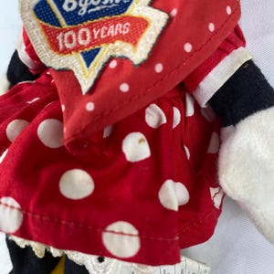 K&ouml;nnte beinhalten: Nahaufnahme einer Vintage-Puppe von OshKosh B'gosh. Die Puppe tr&auml;gt ein rotes Kleid mit wei&szlig;en Punkten und eine rote Jacke. Die Jacke hat einen Aufn&auml;her mit der Aufschrift "100 Years". Die Arme und Beine der Puppe sind schwarz und wei&szlig;.