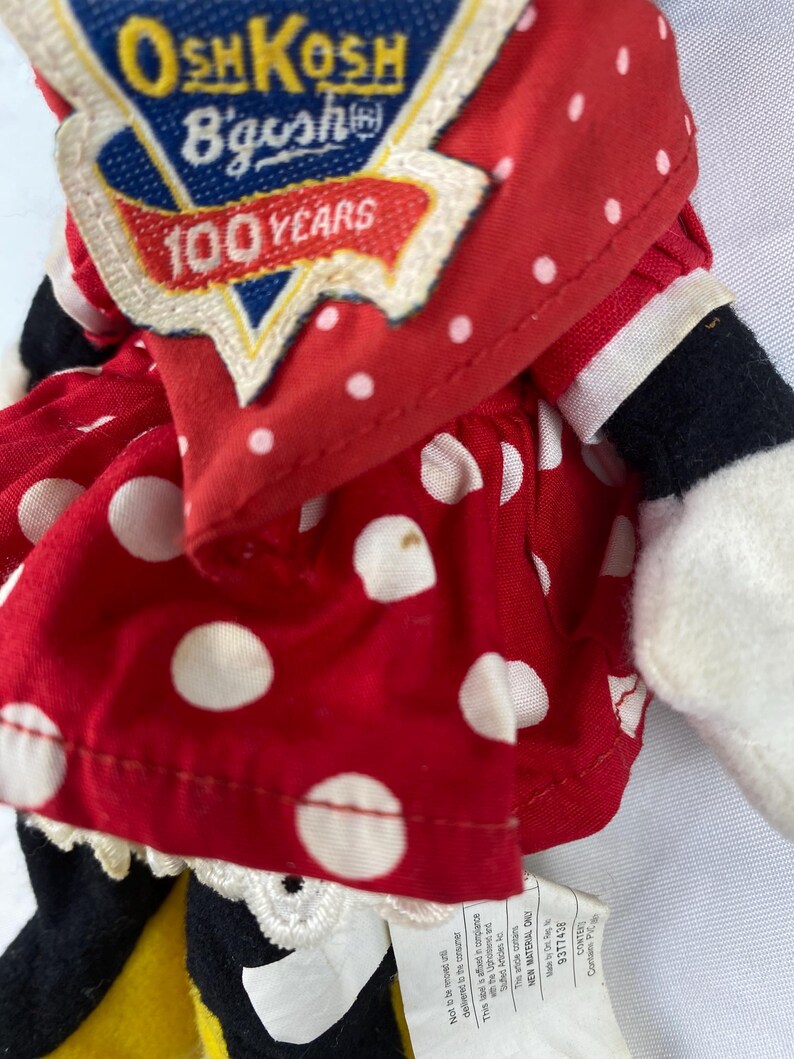 K&ouml;nnte beinhalten: Nahaufnahme einer Vintage-Puppe von OshKosh B'gosh. Die Puppe tr&auml;gt ein rotes Kleid mit wei&szlig;en Punkten und eine rote Jacke. Die Jacke hat einen Aufn&auml;her mit der Aufschrift "100 Years". Die Arme und Beine der Puppe sind schwarz und wei&szlig;.