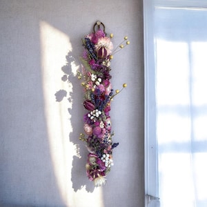 May include: A vertical wall hanging arrangement of dried flowers featuring deep purple, magenta, and pink blossoms mixed with white berries and small yellow buds. The floral display is secured at the top with a circular metal ring for mounting.