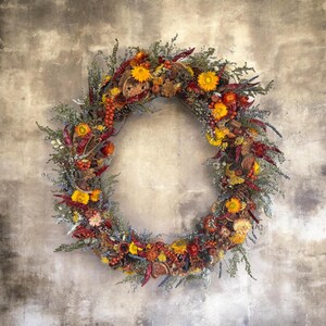 Puede incluir: Una corona circular compuesta por elementos botánicos secos que incluyen girasoles de color amarillo brillante, chiles rojos, bayas naranjas, piñas marrones y varios follajes verdes. El arreglo presenta una mezcla de texturas y tonos otoñales cálidos.
