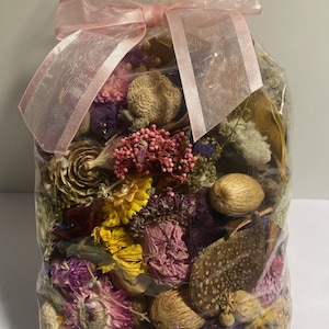 May include: A clear cellophane bag filled with a variety of dried flowers and botanicals. The bag is tied with a sheer pink ribbon. The contents include yellow, purple, and pink flowers, along with seed pods and leaves.