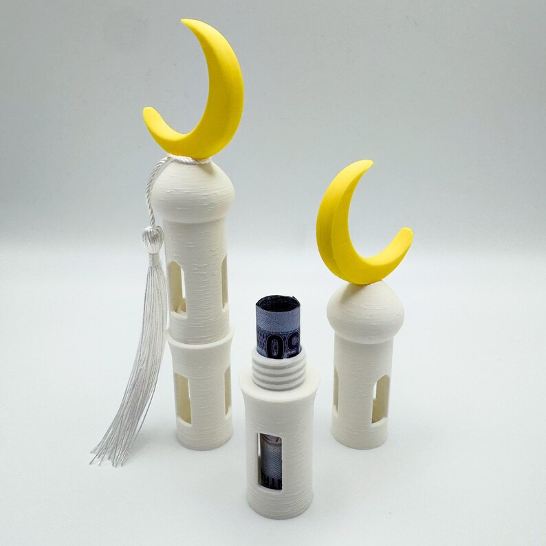 May include: Three white 3D printed mosque-shaped containers with yellow crescent moon tops. The middle container has a rolled-up bill of money inside.