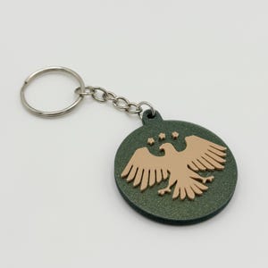 May include: A round keychain with a green background and a tan eagle design. The keychain has a silver chain and ring. The eagle has its wings spread and is centered on the keychain. Three stars are above the eagle.