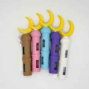 May include: Five decorative, colorful, small, cylindrical containers with a crescent moon on top. The containers are brown, pink, light blue, purple, and white. Each container has a white tassel.