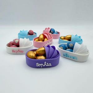 May include: Six small baby shoe-shaped containers filled with chocolate candies. The containers are in various colors, including pink, blue, purple, and white. Each container has a name written on it, including "Sophia", "Oliver", and "Samy".