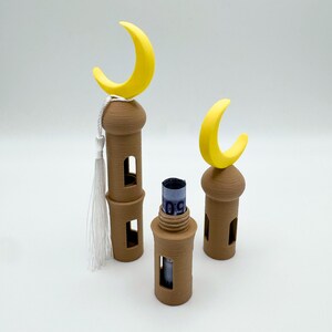 May include: Three brown 3D printed mosque-shaped money boxes with yellow crescent moon tops. The middle box has a rolled-up bill inside.