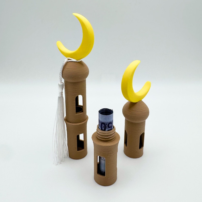 May include: Three brown 3D printed mosque-shaped money boxes with yellow crescent moon tops. The middle box has a rolled-up bill inside.