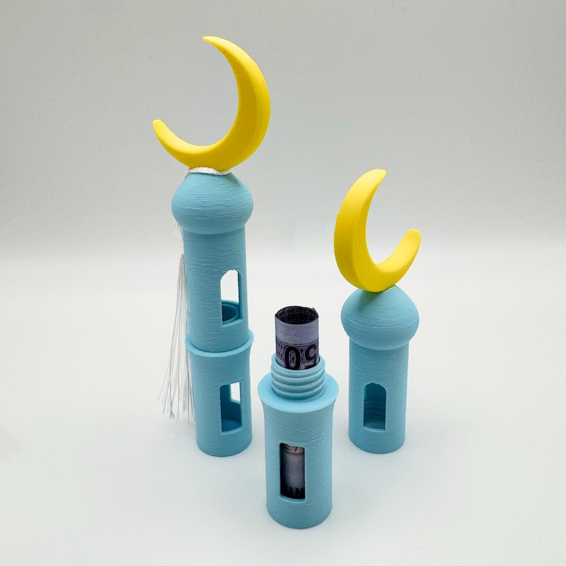 May include: Three blue and yellow 3D printed mosque-shaped containers with a crescent moon on top. The middle container has a rolled-up bill of money inside.
