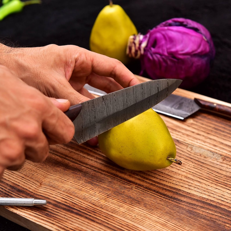 May include: A person's hand holding a black kitchen knife cutting a yellow pear on a wooden cutting board. A purple cabbage and a second yellow pear are in the background.