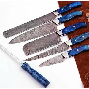 May include: A set of six kitchen knives with patterned steel blades and blue wooden handles. The set includes a sharpening steel. The handles have a marbled blue finish and are riveted to the blades. The knives are displayed on a white surface.